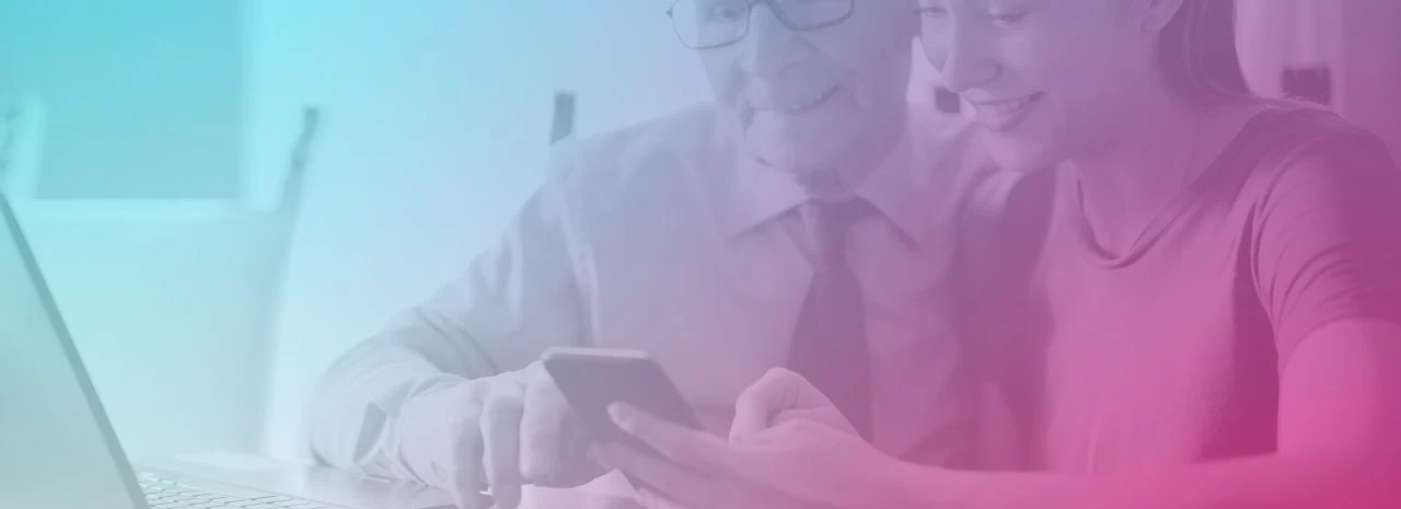 Image of an older man looking at a phone with a younger woman. There is a laptop open in front of them.